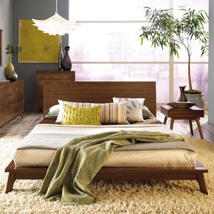 Wooden bed with green and beige bedding, decorative pillows, dresser, and side table in natural lighting.