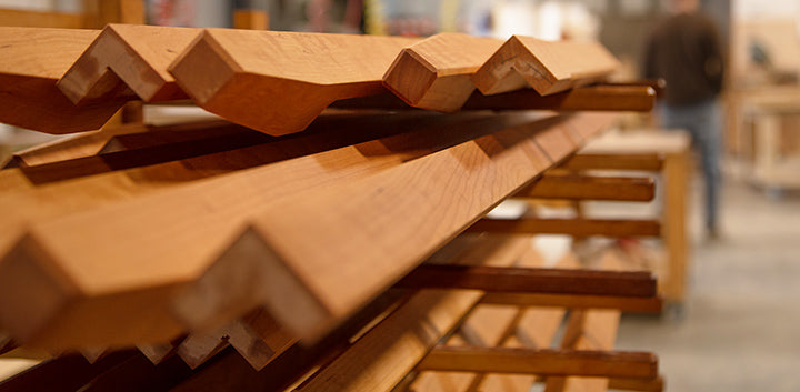 Stacks of wooden planks with notched edges are arranged in a woodworking shop. The background shows an out-of-focus person and workbench.