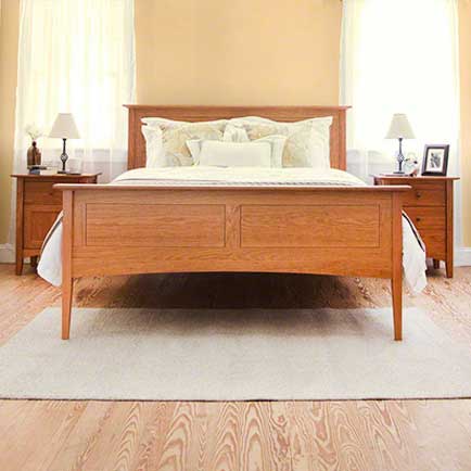 A bedroom with a wooden bed frame, white bedding with pillows, a beige rug, and two matching wooden nightstands on either side. Each nightstand has a lamp and framed picture. The room has wooden floors and light-colored walls with curtains on windows.