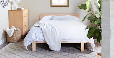 A neatly made bed with white bedding and wooden frame is placed in a bedroom. There is a wooden dresser to the left, a large green plant to the right, and a wicker basket beside the dresser. The room has light-colored walls and a patterned rug on the floor.