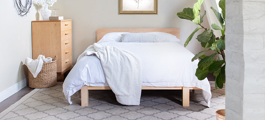 A bedroom with a neatly made bed featuring white bedding and a light wooden frame. A tall plant is to the right of the bed and a wicker basket with a white blanket is placed nearby. A wooden dresser with several drawers is against the back wall.