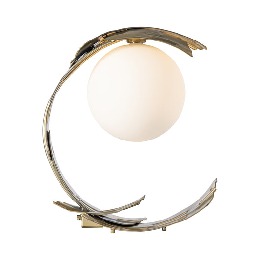 Hubbardton Forge Crest Table Lamp featuring a spherical white light, curved hand-forged steel frame, and Modern Brass finish.