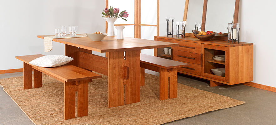 A dining room features a wooden table with two matching benches on a beige rug. A white vase with flowers and various table settings are arranged on the table. A matching wooden sideboard with a mirror, dishes, and a pitcher is in the background.