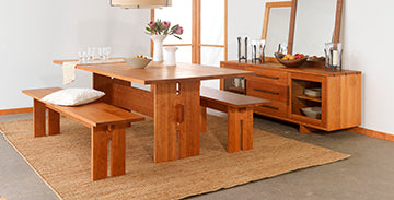 A dining area featuring a wooden table with two matching benches, a wooden sideboard with a mirror above it, and various decorative items including plants, candles, and glassware. The room is bright with natural light and has a jute rug on the floor.