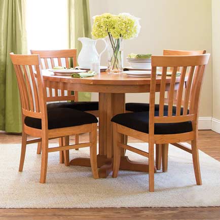 A wooden dining table set with four chairs is placed on a light-colored rug. The table is set with plates, cutlery, and a white pitcher. A vase with yellow flowers is in the center of the table. Green curtains hang by the window in the background.