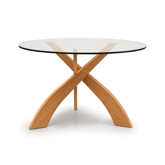 Entwine Round Glass Top Dining Table by Copeland Furniture with intersecting cherry wood legs and a clear glass top.