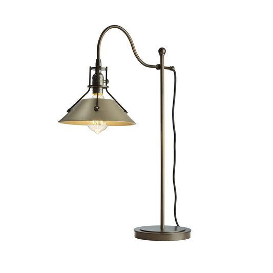 Henry Table Lamp by Hubbardton Forge with bronze finish, curved arm, conical shade, and exposed bulb on a round base creates an industrial aesthetic.