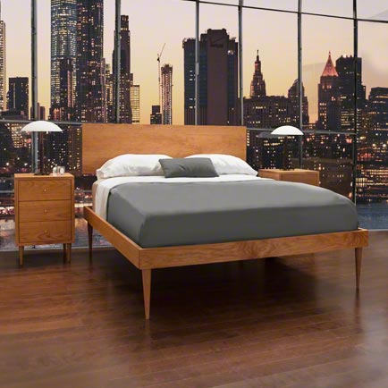 A modern bedroom features a wooden platform bed with a gray comforter and white pillows. Two matching nightstands with lamps flank the bed. A large window behind the bed showcases a city skyline at dusk. The room has a polished wooden floor.