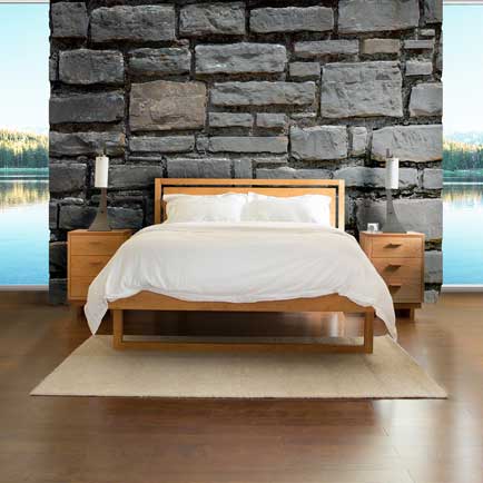 A bedroom with a stone accent wall, a wooden bed frame with white bedding, two small wooden nightstands with candles and lamps on each side, and a tan area rug on a hardwood floor. Large windows behind the bed offer a view of a lake and trees.