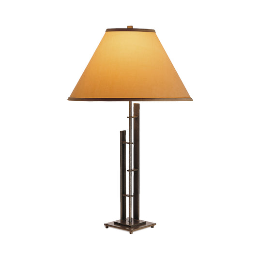 Metra Double #2 Table Lamp by Hubbardton Forge showcases a handcrafted beige conical shade atop a striking metallic geometric base, highlighting exquisite craftsmanship in American furniture design.
