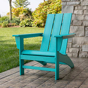 A bright turquoise Adirondack chair is positioned on a stone patio. The chair faces a well-manicured lawn with green bushes and trees in the background. A stone wall is on the right side of the image.