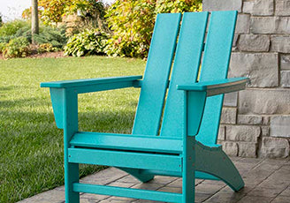 A teal wooden Adirondack chair is placed on a stone patio in a garden setting. The lawn and various green foliage are visible in the background. The chair has a slatted back and wide armrests.