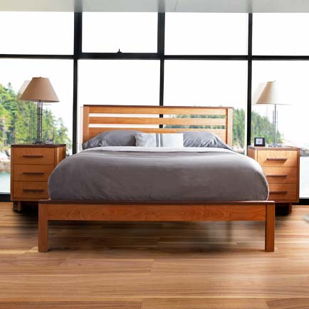 A bedroom with a large wooden bed frame, gray bedding, and two matching wooden nightstands with lamps. The room has floor-to-ceiling windows overlooking a scenic outdoor view. The flooring is wooden. An alarm clock and decorative items are on the nightstands.