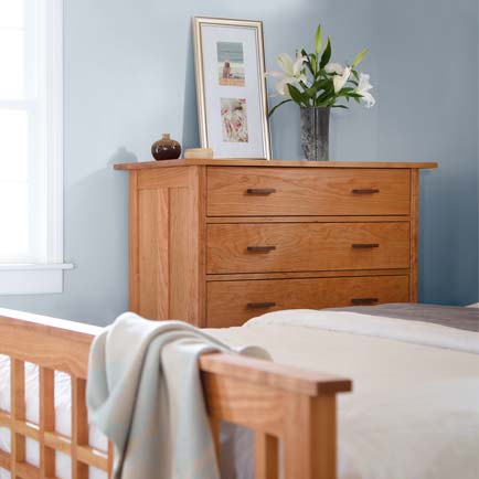 A wooden dresser with three drawers stands against a light blue wall. On top of the dresser are a framed picture, a vase with white flowers, and a small decorative item. To the left is a window, and part of a wooden bed frame is visible in the foreground.