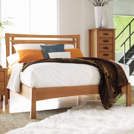 Wooden bed with white sheets and orange pillows, complemented by a decorative throw blanket, alongside a five-drawer dresser.