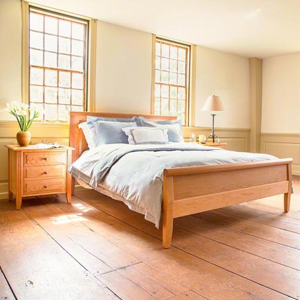 A bedroom with natural light features a wooden bed with a grey duvet and multiple pillows. A wooden nightstand with a potted plant and a lamp is positioned beside the bed. Two large windows above the bed let in sunlight onto the wooden floor.
