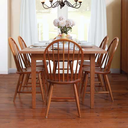A wooden dining table set with six matching chairs is arranged in a bright room with light-colored walls. The table is set with white plates and a vase of white flowers as a centerpiece. A chandelier hangs above the table, and light streams in from a window.