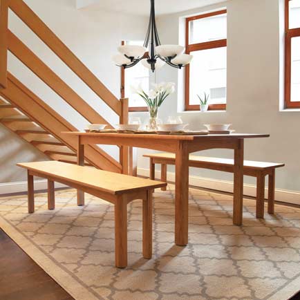 A dining area features a wooden table set with white dinnerware and a vase of white flowers. The seating includes two benches and a chair. A modern chandelier hangs above the table. A patterned rug lies beneath the furniture, and a staircase with wooden railings is visible.